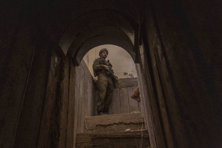 A imagem mostra um soldado em um ambiente escuro, posicionado em uma escada que leva a um espaço mais iluminado. O soldado está de pé, segurando uma arma, e usa um uniforme militar e um capacete. As paredes ao redor são de concreto e parecem desgastadas, com manchas visíveis. A iluminação é baixa, criando um contraste entre a figura do soldado e o fundo.