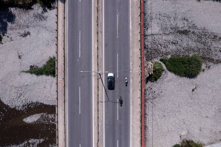 A imagem mostra uma vista aérea de uma estrada com uma ponte. No centro da estrada, há um carro preto e uma motocicleta. Ao redor, é possível ver áreas de vegetação em meio a milhões de peixes mortos