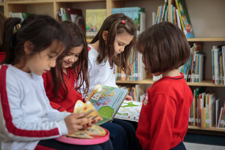 Quatro crianças de idade próxima a seis anos examinam livros em biblioteca. Duas delas usam suéteres vermelhos, e duas estão com blusas brancas.