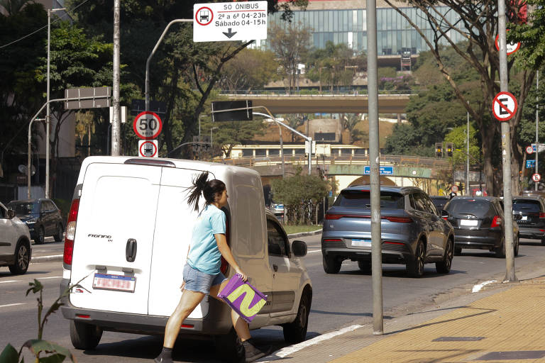 Entregadora de camiseta azul e shorts jeans sobe na calçada segurando uma caixa da empresa Netshoes. Atrás dela, seu carro parado na rua, em frente a uma placa de proibido parar. Outros carros passam no fundo.