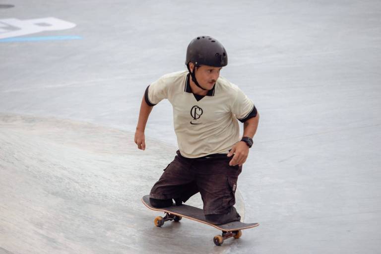 Um skatista está se movendo em uma pista de skate. Ele usa um capacete preto e uma camiseta clara com um logotipo. O skatista está em uma posição inclinada, com um dos joelhos apoiado no skate e o outro pé no chão. O ambiente é uma pista de skate com superfície de concreto.