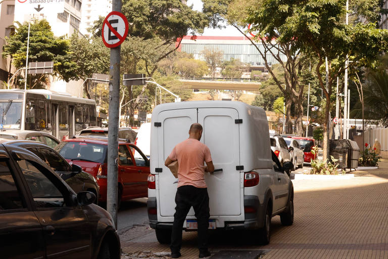 A imagem mostra um homem de costas, vestido com uma camisa clara, abrindo a porta traseira de uma van branca em uma rua urbana. Ao fundo, há árvores e edifícios, além de um sinal de trânsito que indica proibição de parar. Vários carros estão estacionados ao longo da rua.