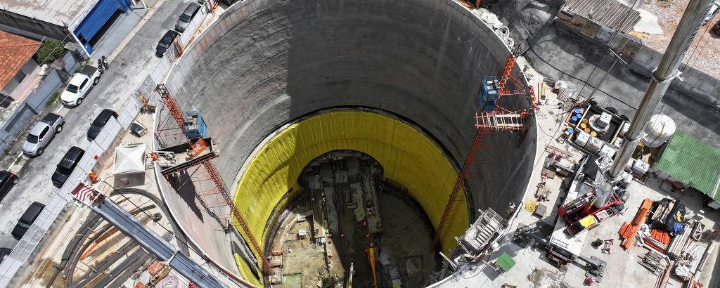 A imagem mostra vista aérea de um poço de construção circular, com paredes de concreto cinza e um fundo amarelo. Ao redor do poço, há várias máquinas e equipamentos de construção, além de veículos estacionados nas proximidades. O ambiente urbano é visível, com casas e ruas ao redor