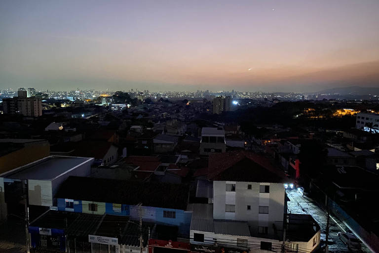 A imagem mostra uma vista panorâmica de uma cidade ao entardecer, com o céu apresentando um gradiente de cores que vai do azul ao laranja. As luzes das casas e edifícios estão acesas, criando um contraste com o céu escuro. Há uma variedade de construções, incluindo casas e prédios, e a imagem captura a sensação de uma área urbana em transição para a noite.