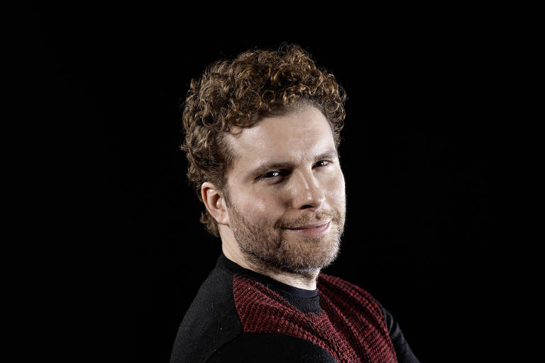 A imagem mostra um homem com cabelo cacheado e barba, olhando para a câmera com um leve sorriso. Ele está vestindo uma camiseta preta com detalhes em vermelho. O fundo da imagem é preto, destacando o rosto e a expressão do homem.