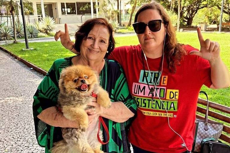 Uma mulher mais velha de vestido verde segura um cachorrinho no colo e tem ao seu lado um outra mulher, de calça azul marinho e camiseta vermelha