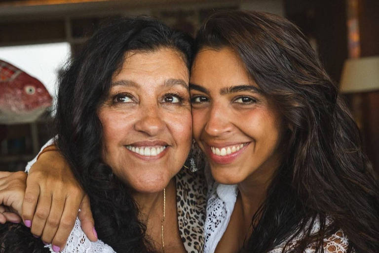 Duas mulheres negras se abraçam e sorriem. A da direita, mais jovem, veste uma blusa rendada branca, de manga comprida, e olha para a foto. A da esquerda, mais velha, está olhando para cima e sorrindo também.