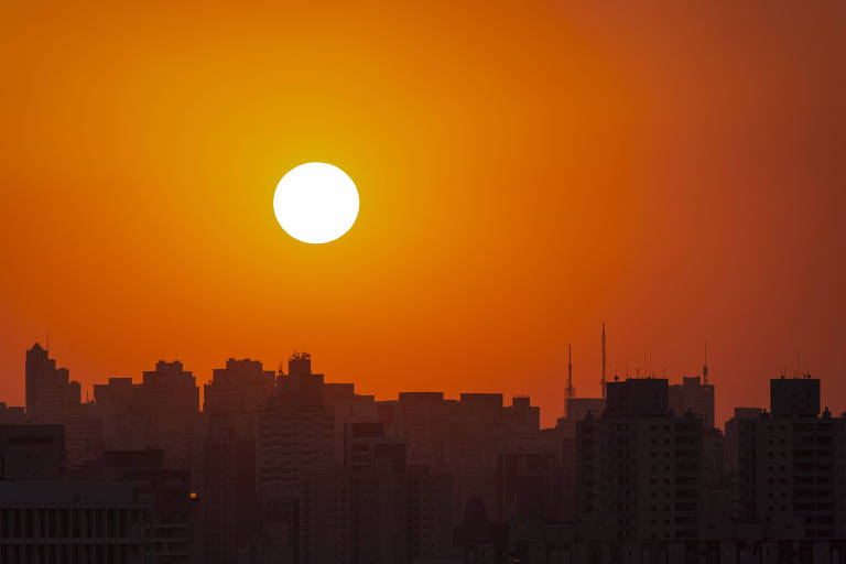 A imagem mostra um pôr do sol, com um grande sol amarelo brilhando no horizonte. A silhueta de uma cidade é visível na parte inferior, com edifícios de diferentes alturas e formas, contra um céu laranja e amarelo.
