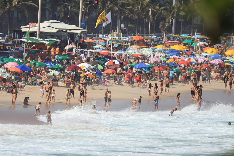 imagem geral mostra banhistas na orla da praia no rio de janeiro; praia lotada com barracas coloridas ao fundo.