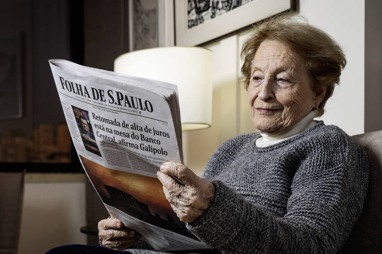 Uma mulher idosa está sentada em uma poltrona, segurando um jornal intitulado 'FOLHA DE S.PAULO'. Ela tem cabelo grisalho e usa um suéter cinza. Ao fundo, há uma luminária acesa e algumas fotos na parede.
