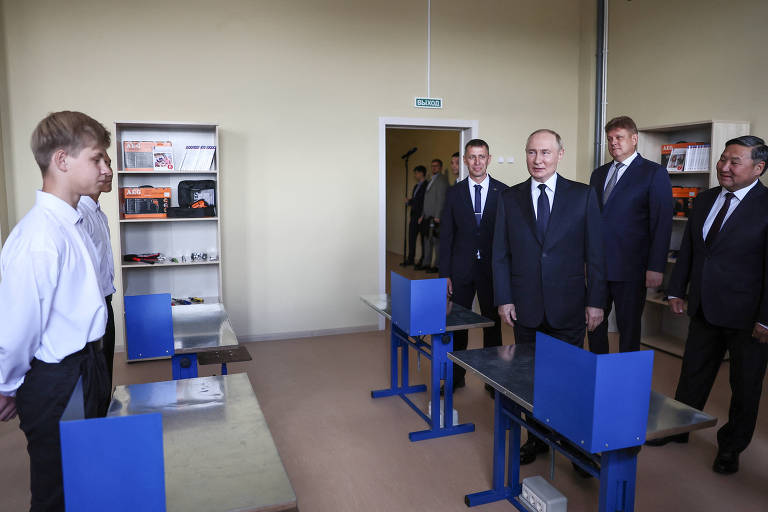 A imagem mostra um grupo de homens em um ambiente interno, possivelmente uma sala de aula ou um centro de treinamento. Um jovem está em pé à esquerda, vestido com uma camisa branca, enquanto cinco homens, incluindo um que parece ser uma figura de destaque, estão à direita, todos vestidos com ternos escuros. Ao fundo, há prateleiras com materiais organizados e mesas azuis na frente.