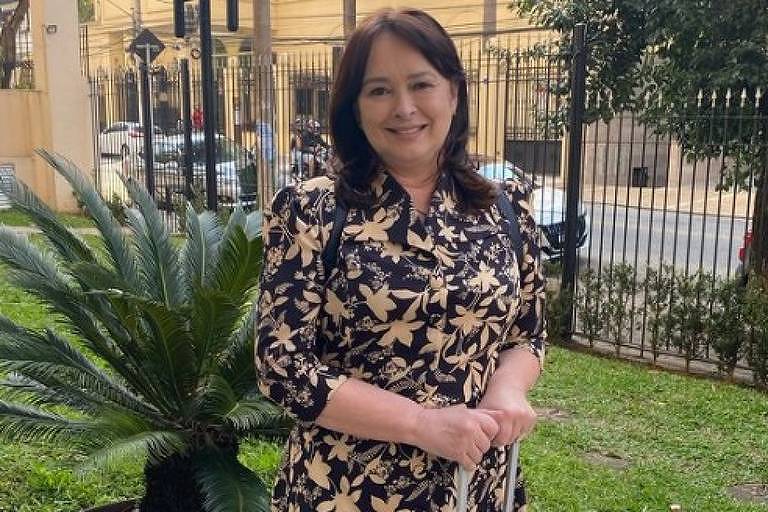 Em foto colorida,  mulher de vestido estampado posa com uma mala em frente ao jardim de um prédio