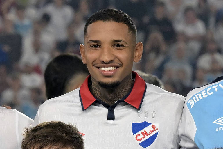 Um jogador de futebol sorri para a câmera, usando uma camisa branca com detalhes em vermelho e azul. O emblema do clube é visível no lado esquerdo do peito, apresentando as letras 'C.N. de F.' e um escudo. O jogador tem tatuagens visíveis no pescoço e um corte de cabelo curto. Ao fundo, há uma multidão desfocada, sugerindo um ambiente de jogo.