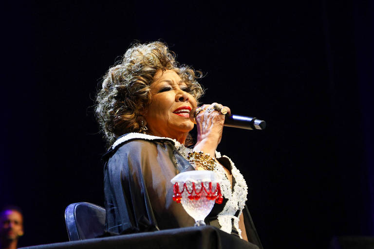 A imagem mostra uma cantora em uma apresentação ao vivo. Ela está segurando um microfone e cantando, com um sorriso no rosto. A cantora tem cabelo cacheado e usa uma roupa escura com detalhes claros. À sua frente, há um copo decorativo. O fundo é escuro, sugerindo um ambiente de palco.