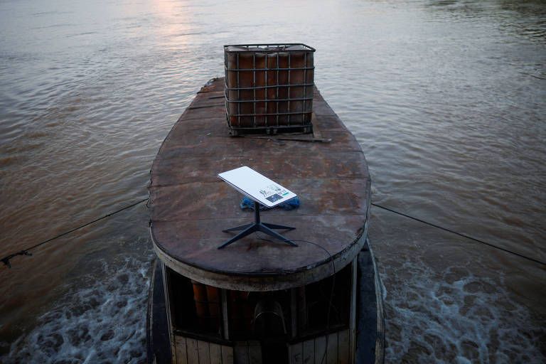 A imagem mostra a parte superior de um barco flutuando em água. No centro, há um equipamento da Starlink parecido com um tablet, a superfície é branca com pernas pretas, e ao fundo, uma cesta de material em cima do barco. A água ao redor reflete a luz do sol, criando um ambiente tranquilo.