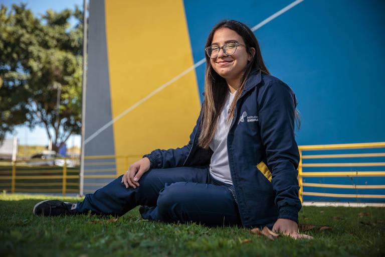 Uma jovem está sentada no gramado, vestindo um conjunto de roupas esportivas escuras. Ela usa óculos e tem cabelo longo e liso. Ao fundo, há uma parede colorida com tons de azul e amarelo, e algumas árvores visíveis na parte superior da imagem.