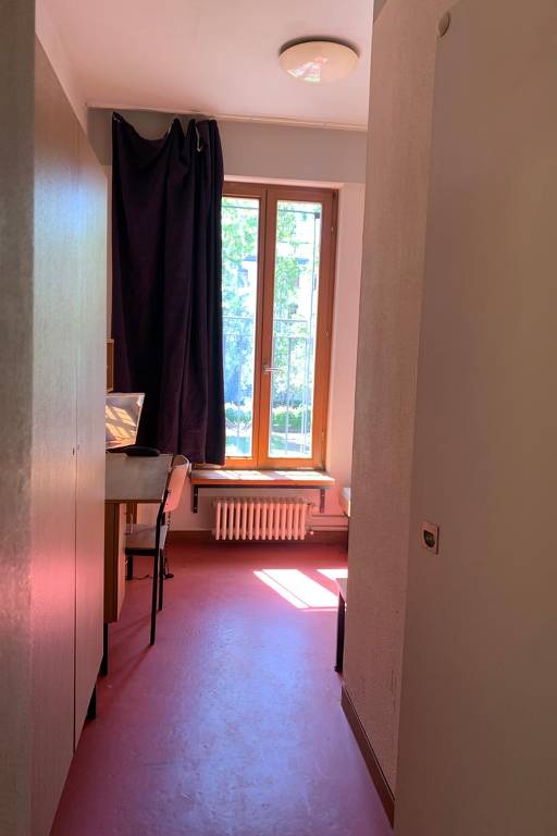 A imagem mostra a entrada de um quarto com uma parede clara à esquerda e uma janela à direita, onde há cortinas escuras. A luz do sol entra pela janela, iluminando o chão de cor rosa. Há uma mesa com uma cadeira à esquerda e um aquecedor sob a janela. O ambiente parece ser bem iluminado e arejado.