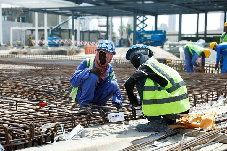 A imagem mostra dois trabalhadores da construção civil em um canteiro de obras. Um dos trabalhadores está agachado, usando um macacão azul e um lenço cobrindo parte do rosto, enquanto o outro está de joelhos, vestindo uma camiseta preta e um colete refletivo amarelo. Ao fundo, há outros trabalhadores e estruturas de metal em construção, com materiais de construção espalhados pelo chão.
