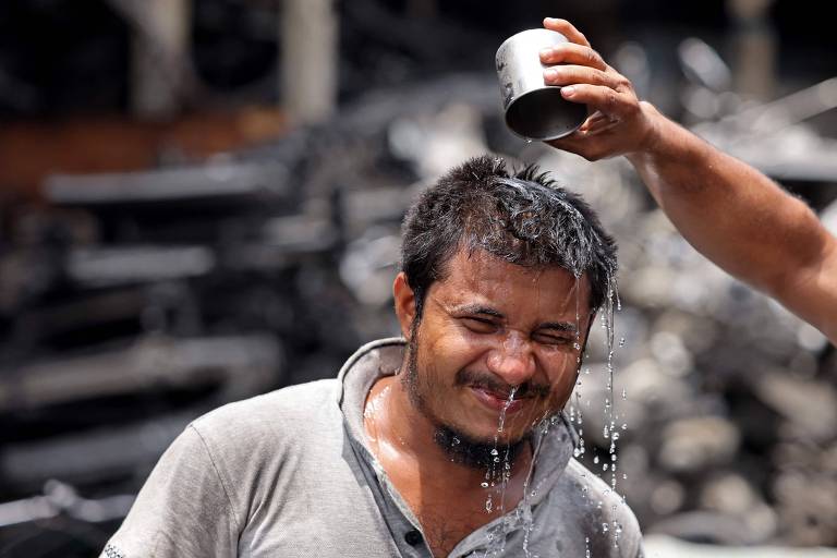 Um homem está sorrindo enquanto recebe água derramada sobre sua cabeça de um recipiente metálico. Ele está com a camisa cinza e seu cabelo está molhado, com gotas de água escorrendo pelo seu rosto. Ao fundo, há uma cena desfocada de objetos escuros, possivelmente sucata.
