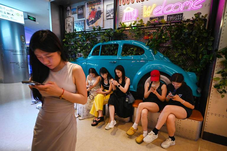 A imagem mostra um grupo de cinco mulheres sentadas em um banco, todas olhando para seus celulares. Uma mulher em pé à frente está também usando seu celular. Ao fundo, há um carro azul decorativo e plantas. O ambiente parece ser um espaço público ou comercial.