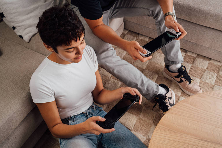 A imagem mostra duas pessoas sentadas em um sofá, jogando videogame. Uma pessoa, com cabelo curto e escuro, usa uma camiseta branca e segura um controle de videogame. A outra pessoa, parcialmente visível, está segurando um controle semelhante. O ambiente é aconchegante, com um piso de madeira e uma mesa de centro ao lado.