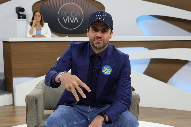 Pablo Marçal está sentado em um sofá em um estúdio de televisão, vestindo um blazer escuro e uma camiseta. Ele está fazendo um gesto com a mão. Ao fundo, uma mulher está em pé, aparentemente apresentando algo, com um cenário moderno e uma tela visível.