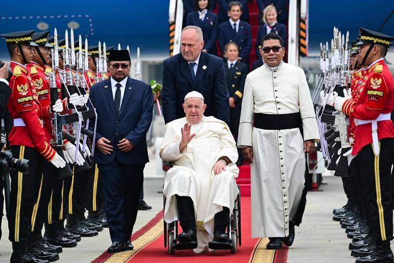 A imagem mostra uma cerimônia de recepção ao Papa, que está em uma cadeira de rodas, sendo acompanhado por dois homens. Ele está vestido com uma túnica branca. Ao redor, há uma formação de guardas em uniformes cerimoniais, segurando espadas. Ao fundo, é possível ver um avião e outras pessoas assistindo ao evento.