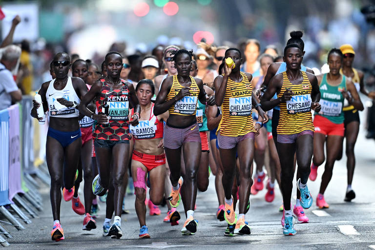 A imagem mostra um grupo de mulheres competindo em uma corrida. Elas estão vestidas com trajes de atletismo, com algumas usando uniformes de diferentes países, como Kenya e Uganda. O cenário é urbano, com espectadores ao fundo e uma atmosfera de competição. As corredoras estão em movimento, algumas com expressões de concentração.