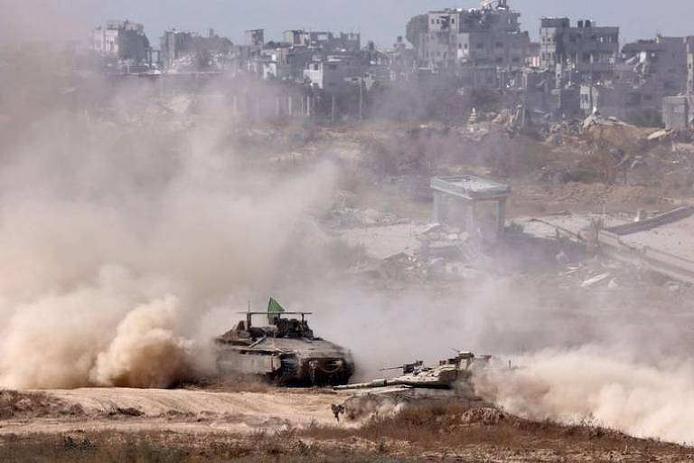 A imagem mostra um tanque de guerra em movimento em uma área de combate, levantando poeira. Ao fundo, há edifícios danificados e em ruínas, com fumaça e poeira no ar, indicando um ambiente de conflito. O cenário é desolador, com sinais de destruição visíveis.