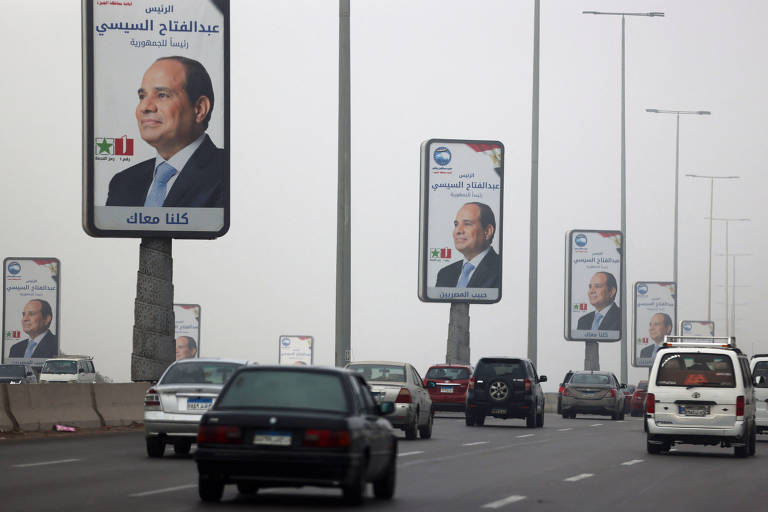 A imagem mostra uma estrada com vários painéis publicitários ao longo dela. Os painéis exibem a imagem de um homem em um terno, com um fundo claro e texto em árabe. Há também símbolos de bandeiras. Vários carros estão circulando na estrada, e o ambiente parece nublado.