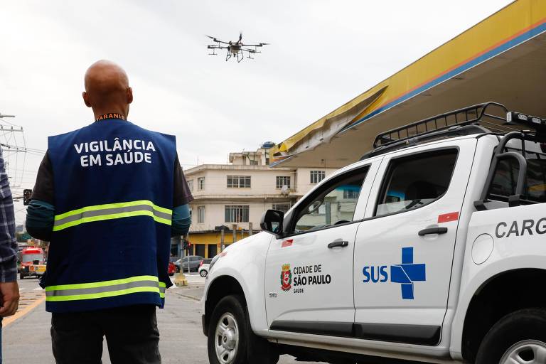 A imagem mostra um homem de costas, usando um colete azul com as palavras 'VIGILÂNCIA EM SAÚDE' em destaque. Acima dele, um drone está voando. À direita, há uma caminhonete branca com a inscrição 'CIDADE DE SÃO PAULO' e o logotipo do SUS. O fundo apresenta um edifício e um céu nublado.