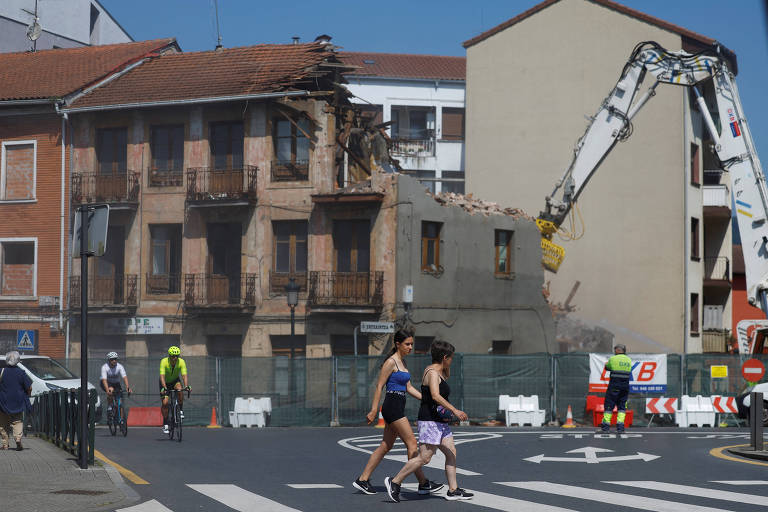 A imagem mostra um edifício em processo de demolição, com parte da estrutura já derrubada. No primeiro plano, duas mulheres estão atravessando a rua, enquanto um ciclista passa ao lado. Ao fundo, uma máquina de demolição está operando, e há uma área cercada com cones de sinalização. O céu está claro e ensolarado.
