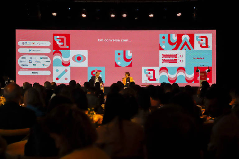 A imagem mostra um evento com um grande painel de fundo em cores vibrantes, onde está escrito 'Em conversa com...'. No palco, duas pessoas estão sentadas, uma delas é um homem e a outra uma mulher, ambos participando de uma discussão. O público está visível em primeiro plano, com mesas e iluminação suave. O painel exibe logotipos de patrocinadores e informações sobre o evento, incluindo 'Fórum Brasileiro de Filantropos e Investidores Sociais'.