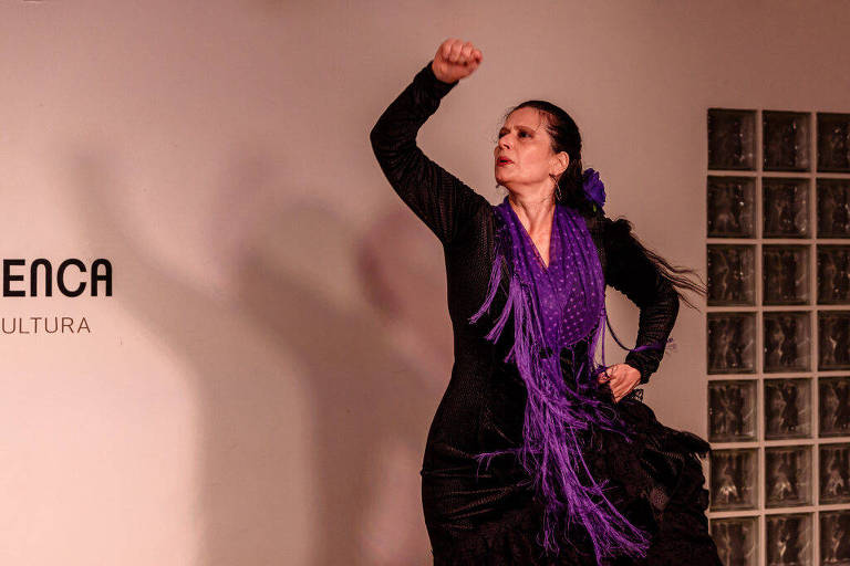 Uma dançarina de flamenco está em uma apresentação, usando um vestido preto e um xale roxo. Ela levanta o braço direito com um gesto expressivo, enquanto seu cabelo longo e escuro está solto. Ao fundo, há uma parede com o texto 'FLAMENCA' e 'CULTURA'.