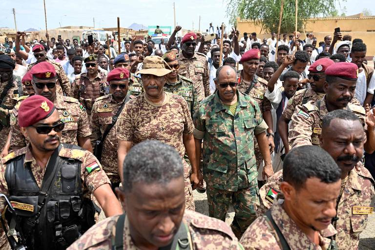 A imagem mostra um grande grupo de pessoas, incluindo militares e civis, em uma área aberta. Os militares estão vestidos com uniformes camuflados e bonés vermelhos, enquanto alguns civis estão em roupas casuais. Há uma atmosfera de agitação, com pessoas levantando os braços e demonstrando apoio ou protesto. Ao fundo, é possível ver uma construção e árvores