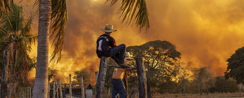 A imagem mostra um homem sentado em uma cerca de madeira, usando um chapéu de palha. Ele observa um incêndio ao fundo, onde há fumaça densa e um céu alaranjado. A cena é em uma área rural, com algumas palmeiras e árvores ao redor, e o solo parece seco e árido.