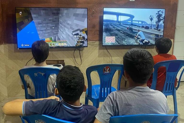 A imagem mostra uma sala de jogos onde quatro crianças estão sentadas em cadeiras plásticas azuis, assistindo a dois televisores. À esquerda, um jogo de Minecraft está sendo exibido, enquanto à direita, um jogo de Grand Theft Auto é visível. As paredes estão decoradas com pôsteres de vários jogos. Há também um aviso na parede com informações sobre preços de locação de consoles.