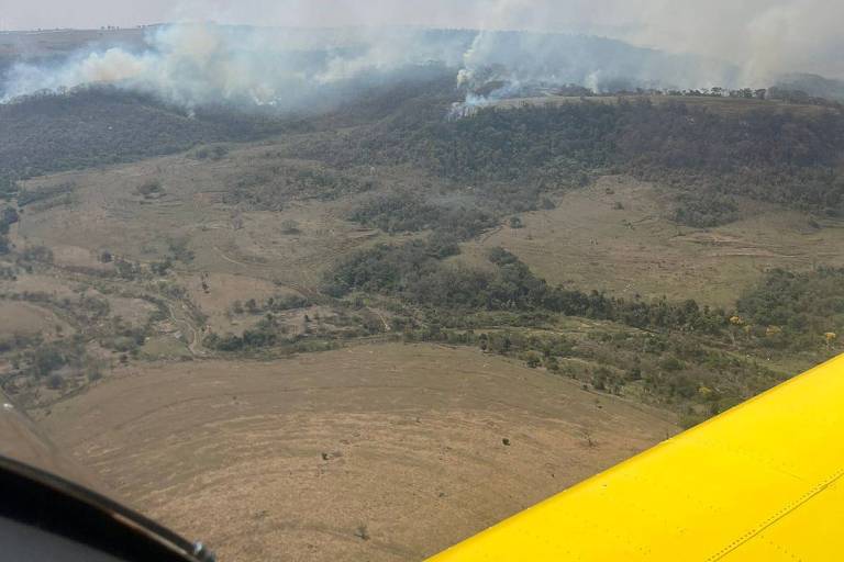 Vista aérea de uma área afetada por incêndio florestal. Há fumaça se elevando de uma região montanhosa ao fundo, enquanto o primeiro plano apresenta uma parte da asa de um avião amarelo. O terreno abaixo é predominantemente seco e árido, com algumas áreas verdes visíveis