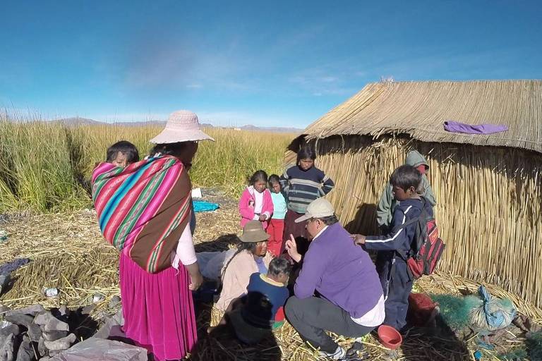 A imagem mostra uma comunidade, com várias pessoas interagindo. Algumas estão vestidas com roupas tradicionais coloridas, enquanto outras estão agachadas ou sentadas no chão. Ao fundo, há uma construção simples feita de palha e um céu azul claro. A cena retrata um ambiente rural e comunitário.