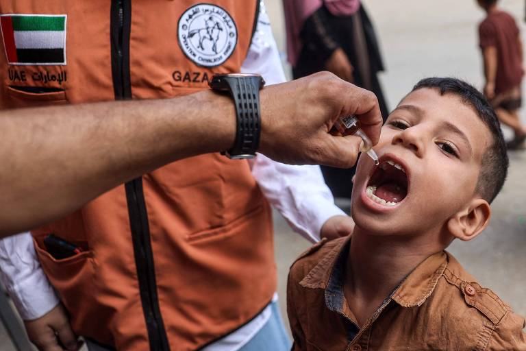 A imagem mostra um menino com a boca aberta, recebendo um medicamento de um adulto que está usando uma camisa com um colete laranja. O colete tem um emblema que parece ser de uma organização de saúde. O fundo apresenta outras pessoas, mas não estão claramente visíveis.