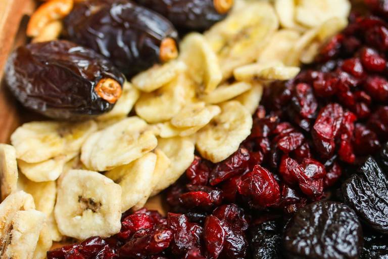 A imagem mostra uma variedade de frutas secas e nozes dispostas em uma bandeja de madeira. À esquerda, há tâmaras escuras e fatias de banana desidratada. No centro, estão cranberries vermelhas e secas. À direita, há ameixas secas escuras e nozes pecan marrons.