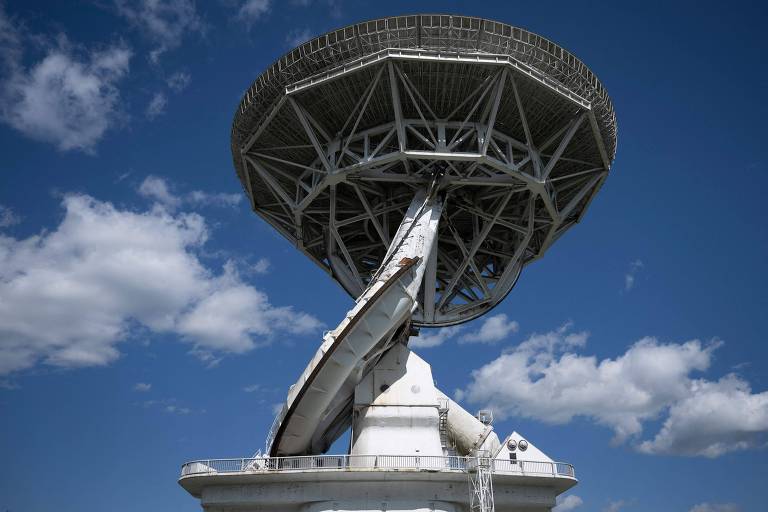 A imagem mostra uma grande estrutura de radar com uma base robusta e uma parte superior em forma de disco, posicionada contra um céu azul com algumas nuvens brancas. A estrutura é predominantemente branca e possui detalhes em metal.
