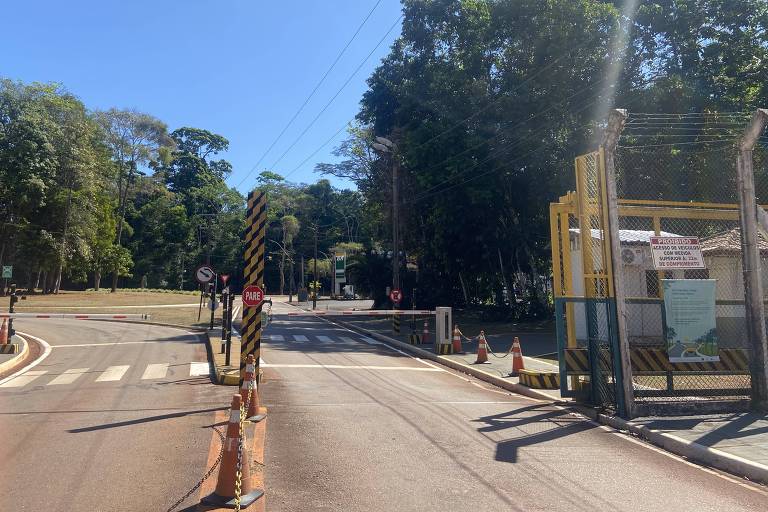 A imagem mostra a entrada de um parque em um dia ensolarado. À esquerda, há uma estrada que se curva, e à direita, uma estrutura amarela que parece ser um portão de entrada. O ambiente é cercado por árvores verdes e um céu azul claro. Não há pessoas visíveis na imagem.