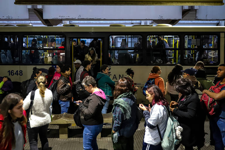 A imagem mostra uma estação de ônibus movimentada, com várias pessoas esperando para embarcar. Algumas estão em pé, enquanto outras estão sentadas em bancos. A maioria das pessoas está olhando para seus celulares. Um ônibus está estacionado ao fundo, com algumas pessoas descendo dele. O ambiente é urbano e parece ser um horário de pico.