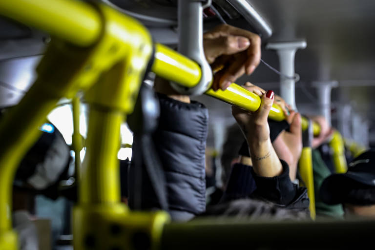 A imagem mostra várias mãos segurando barras amarelas em um ônibus. As mãos são de diferentes pessoas, algumas com unhas pintadas e outras com luvas. O fundo é desfocado, mas é possível ver parte do interior do ônibus, com assentos e outros passageiros.