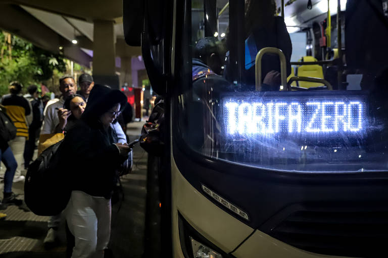 A imagem mostra um ônibus com a inscrição 'TARIFA ZERO' iluminada na frente. Algumas pessoas estão esperando para embarcar, enquanto uma mulher à esquerda está usando um celular. O ambiente parece ser urbano, com iluminação noturna e outras pessoas ao fundo.