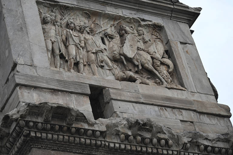 A imagem mostra um alto-relevo esculpido em uma estrutura de pedra, representando figuras humanas em trajes antigos, possivelmente soldados e líderes. As figuras estão em uma cena de batalha ou celebração, com armas e armaduras visíveis. O alto-relevo apresenta detalhes elaborados, mas algumas partes da escultura estão desgastadas ou danificadas.