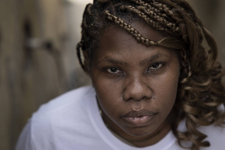 A imagem mostra uma mulher negra com tranças, olhando diretamente para a câmera. Ela tem uma expressão séria e está vestindo uma camiseta branca. O fundo é desfocado, sugerindo um ambiente urbano.