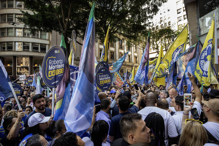 Pessoas aglomeradas no meio de uma rua, entre prédios antigos e árvores, segurando bandeiras com cores azul e amarela