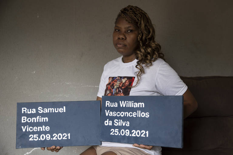 Uma mulher negra sentada em um sofá, segurando duas placas. A placa à esquerda diz 'Rua Samuel Bonfim Vicente 25.09.2021' e a placa à direita diz 'Rua William Vasconcellos da Silva 25.09.2021'. A mulher está usando uma camiseta branca com uma imagem colorida e tem cabelo cacheado e solto.